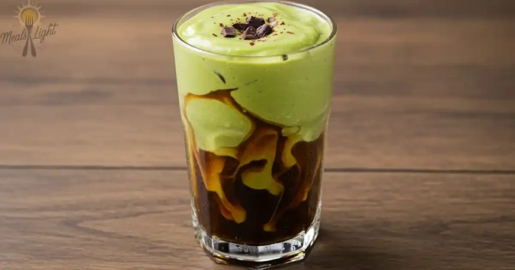 A glass of creamy avocado coffee float with vibrant green avocado cream layered over rich black coffee, topped with chocolate shavings, placed on a wooden table.