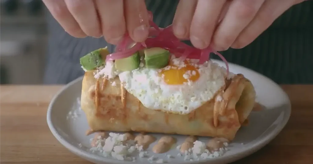 A golden-brown breakfast burrito topped with a fried egg, avocado, crumbled cheese, and pickled red onions.