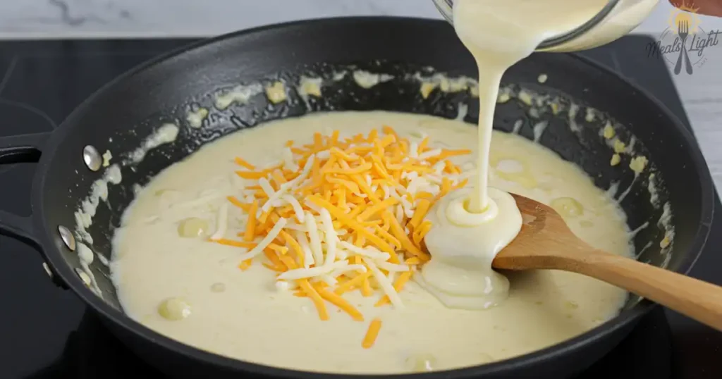 Cheese sauce being poured into a pan with shredded cheese on top, stirred with a wooden spoon.